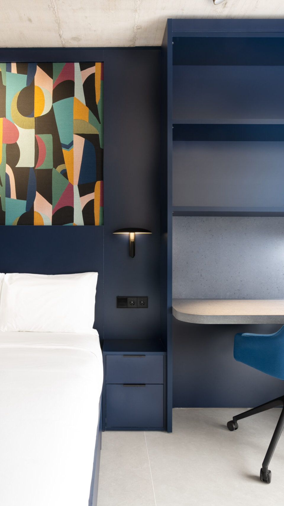 Cama blanca junto a un escritorio empotrado y una silla azul, con arte abstracto en la pared.