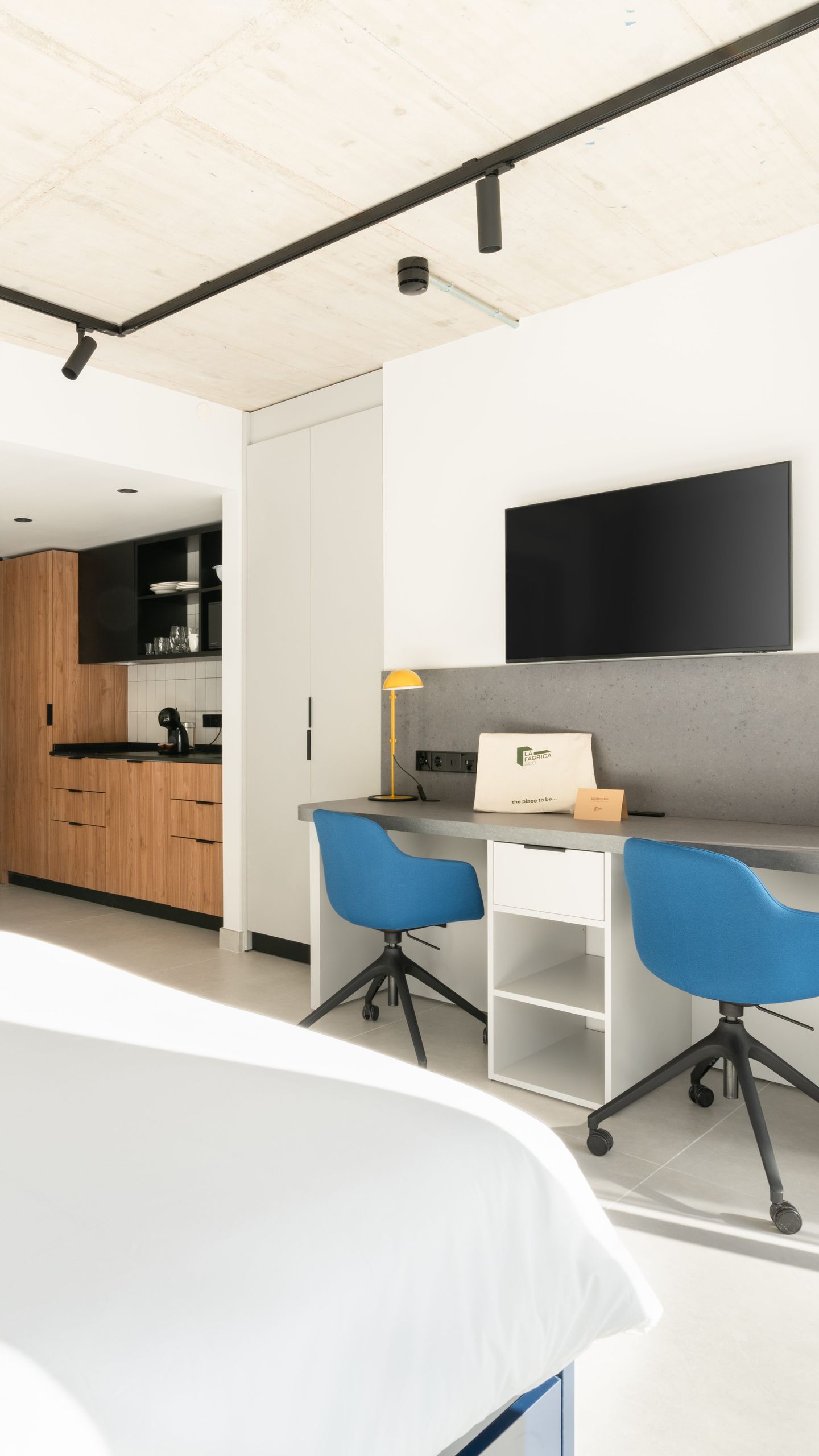 Habitación moderna con escritorio, dos sillas azules, televisor y zona de cocina.