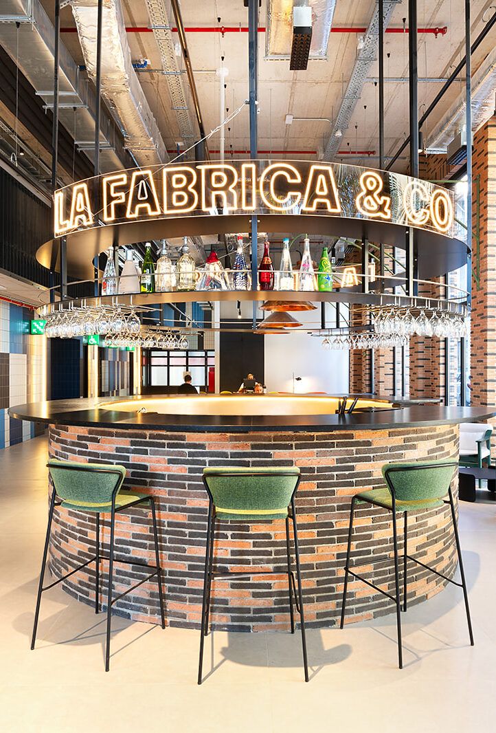 Bar con barra circular de ladrillo, taburetes verdes y letrero neón «LA FABRICA & CO».