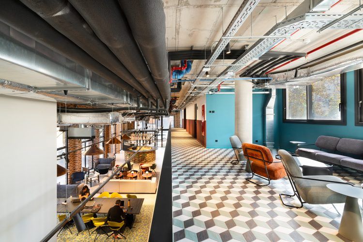 Interior de oficina de planta abierta con suelo estampado, servicios del techo a la vista y zona de lounge con asientos.
