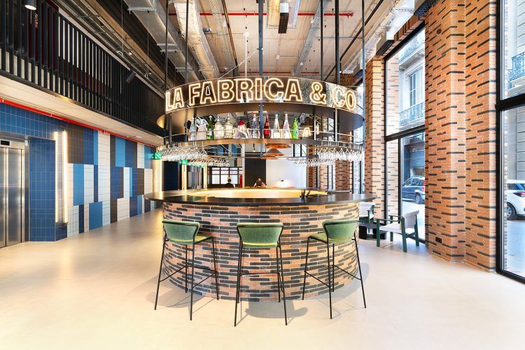 Interior de bar con mostrador circular, copas colgantes y un letrero de LA FABRICA & CO.