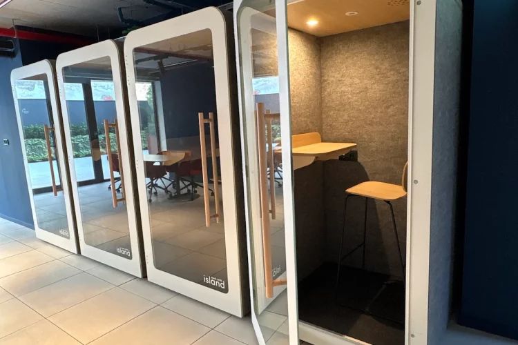 Cabinas telefónicas de vidrio con mesas y taburetes en un espacio de oficina moderno.